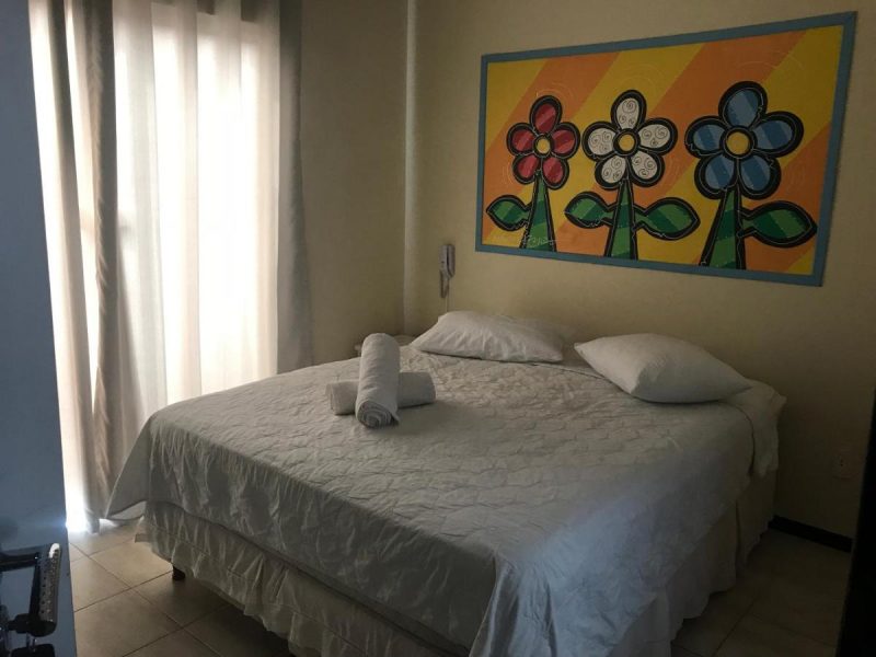 Foto de um quarto do Shallom Hotel, em Balneário Camboriú. Na foto, aparece uma cama de casal com lençol e travesseiros brancos, um quadro com flores coloridas na parede e a cortina em tom claro. 