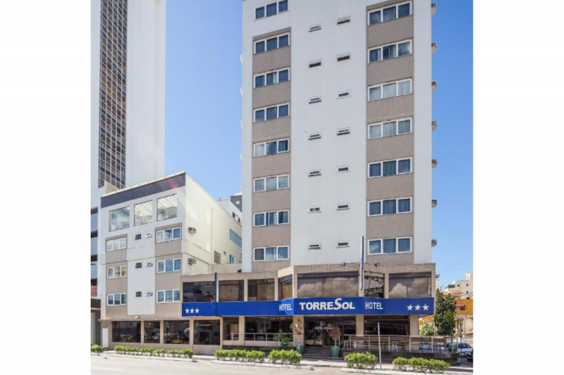 Fachada do Torre Sol, um hotel barato em Balneário Camboriú. O prédio do hotel é alto, em tons claros. No térreo, na entrada do hotel, há uma faixa azul onde está escrito o nome do empreendimento em letras brancas.