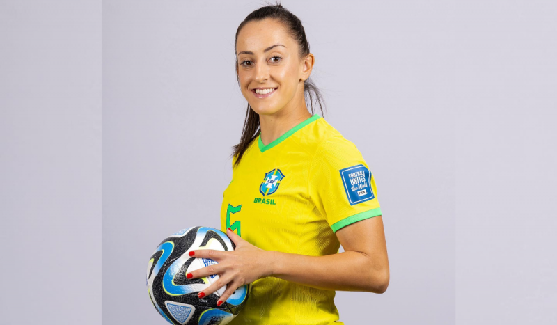 Mulher branca posa de perfil segurando uma bola de futebol e usando uma camisa amarela da seleção brasileira. Foto mostra Luana Bertolucci, jogadora que revelou ter Linfoma de Hodgkin
