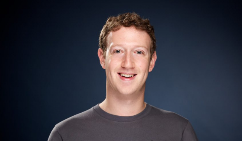 Mark Zuckerberg - Foto mostra homem branco de olhos azuis sorrindo em frente a fundo azul usando camisa cinza