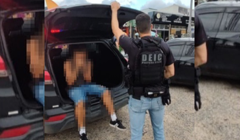 Homem sendo colocado em camburão de carro de polícia enquanto delegado posa de costas para a foto segurando a porta do carro, para ilustrar matéria sobre motorista de aplicativo preso
