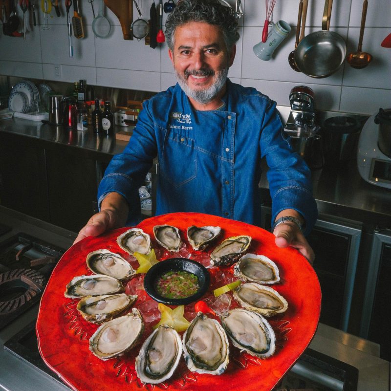 Foto do proprietário do Restaurante Ostradamus, em Florianópolis. Ele é um homem branco, de barba e cabelos grisalhos. Veste uma blusa de manga comprida azul e segura, com os braços esticados, um prato vermelho com 13 unidades de ostras. Ao fundo aparecem utensílios de cozinha. 