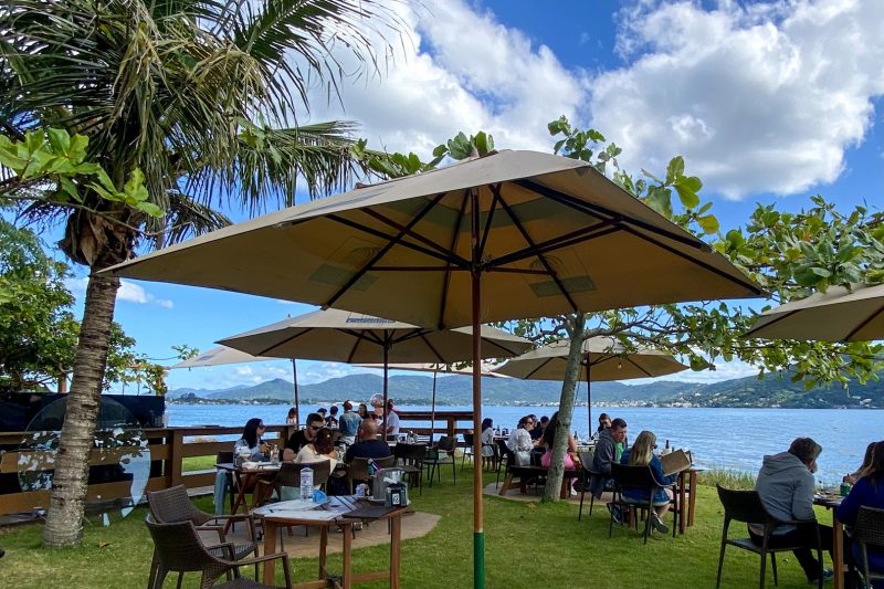 Foto do restaurante Pontas das Caranhas, que serve ostras em Florianópolis. A foto mostra uma área externa gramada, como guarda-sóis, mesas e cadeiras, onde estão sentadas pessoas, de frente para a Lagoa da Conceição. 