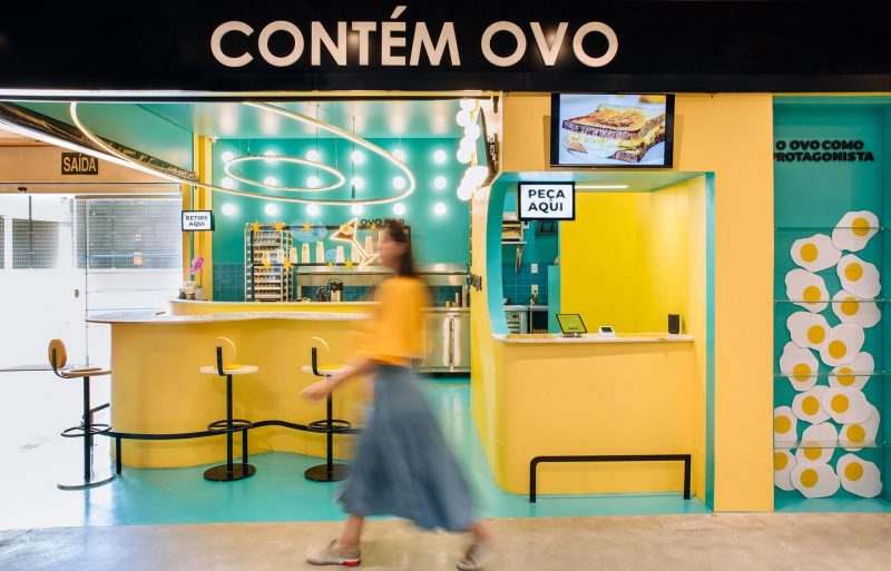 Fachada do restaurante Contém Ovo, dentro do Mercadão de Coqueiros. Todo o mobiliário é verde e amarelo, com desenhos de ovos no lado direito da entrada do estabelecimento. Uma mulher passa pela frente da fachada e sua imagem está desfocada. 