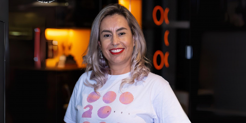 Na imagem vemos Samanta sorrindo para a foto, ela é loira e está com o cabelo solto e uma blusa branca.