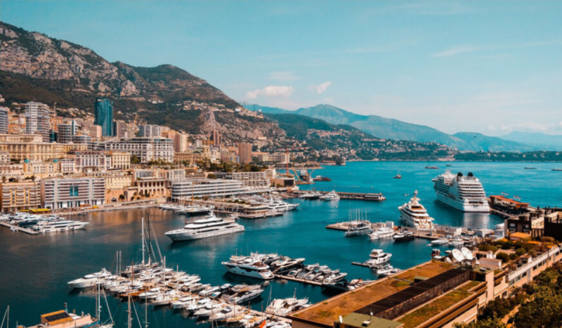 Foto mostra cruzeiros e barcos pequenos atracados em decks. Na imagem também é possível ver o mar azul, montanhas e prédios ao fundo