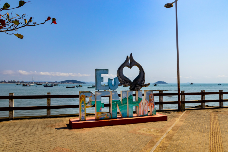 Foto de uma escultura escrito Eu amo Penha. A palavra amo é representada por dois peixes cuja posição lembra o formato de um coração. Ao fundo aparece o mar com alguns barcos e no canto superior esquerdo alguns galhos e folhagens de uma árvore. 