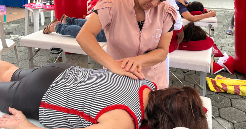 na foto, uma mulher está deitada de bruços enquanto outra mulher apoia duas mãos na sua coluna em forma de massage; ação faz parte do BG nos Bairros