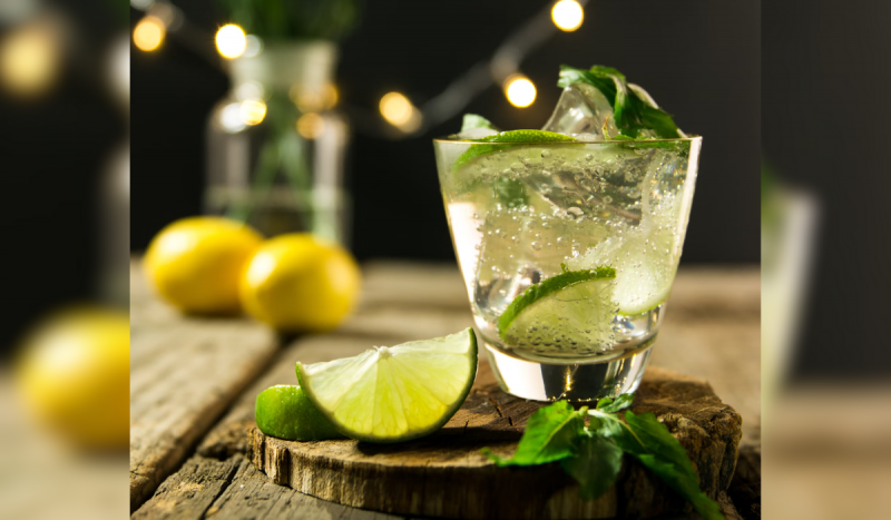 Capirinha em copo transparente. Copo está em cima de roda de madeira com limão cortado ao lado e folhas de hortelã