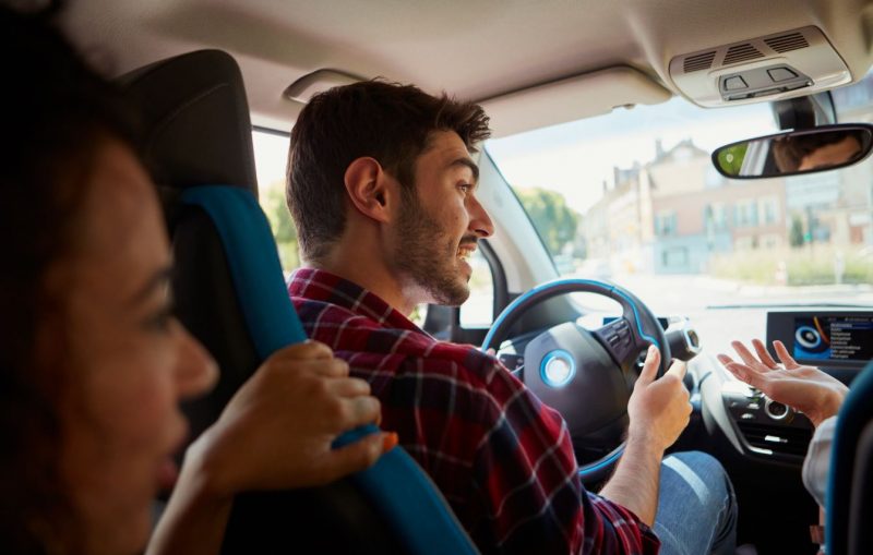 Foto ilustrativa do app de viagem Blablacar. A foto é de dentro de um carro. Um homem de cabelos curtos castanhos, barba serrada e camisa xadrez dirige. Ele olha sorrindo para a passageira que está ao seu lado, da qual aparece apenas a mão. Uma outra passageira está sentada atrás e se apoio no banco do motorista, numa posição que demostra estar conversando com os demais. Ela aparece desfocada na foto. É uma mulher jovem, de cabelos pretos. 