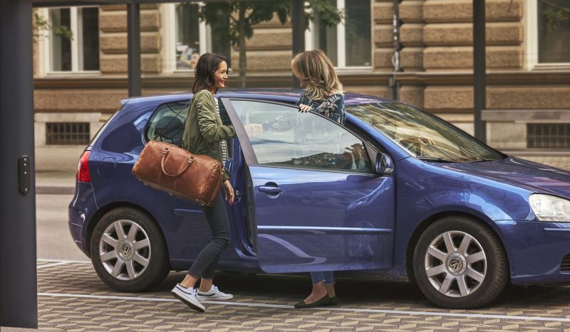 Foto ilustrativa do app de viagem Blablacar. No centro da foto, duas mulheres - uma morena com uma mala no ombro e uma loira - se cumprimentam na porta de um carro azul, parada à beira de uma calçada. 