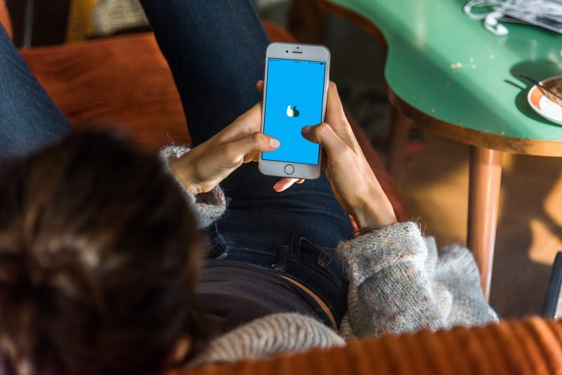 Foto de uma moça de cabelos castanhos, blusa preta e calça jeans azul sentada em um sofá. Ela segura um celular nas mãos e abre o app de viagem Blablacar. A logomarca do app aparece na tela. 