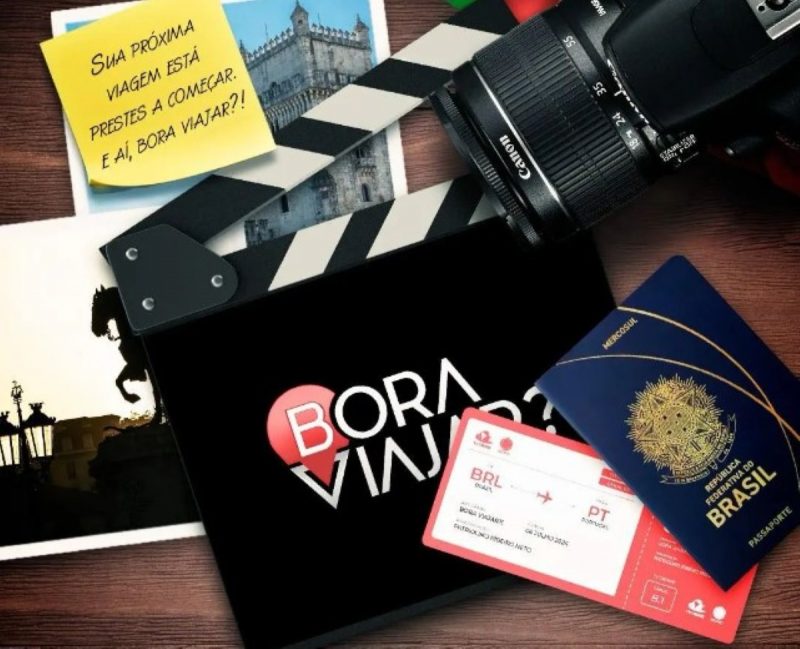 Na imagem aparece uma mesa uma claquete do programa Bora Viajar com um post it, passaporte, fotos e câmera fotográfica e passagem