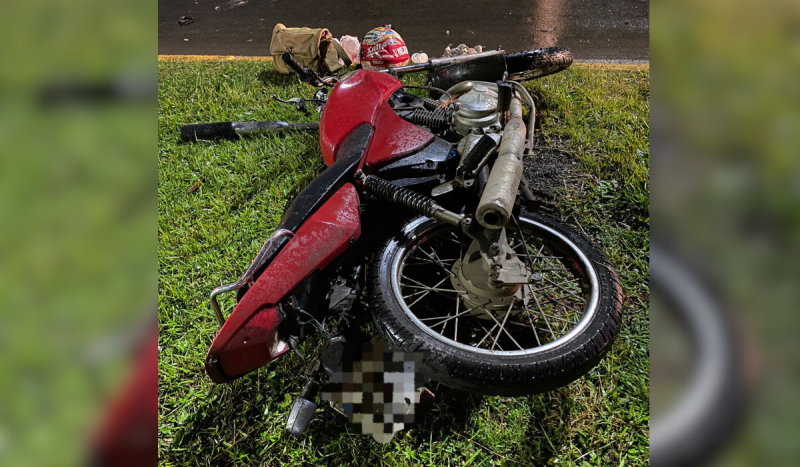 Foto mostra motocicleta vermelha jogada no chão após acidente de trânsito. Veículo está sobre gramado e ao fundo é possível ver uma parte da pista