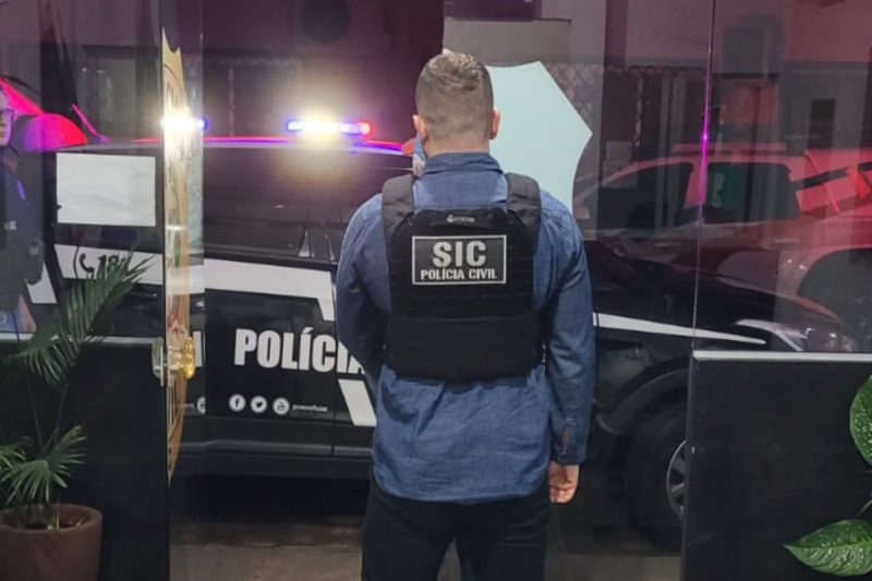 homem foi preso pela polícia civil por oferecer dinheiro a crianças em troca de favores sexuais; na imagem um policial civil de costas com o colete preto escrito SIC Polícia Civil; O policial está com uma camisa de manga longa azul e uma calça preta e tem o cabelo raspado atrás. Ao fundo, um carro da Polícia Civil de SC com as sirenes ligadas