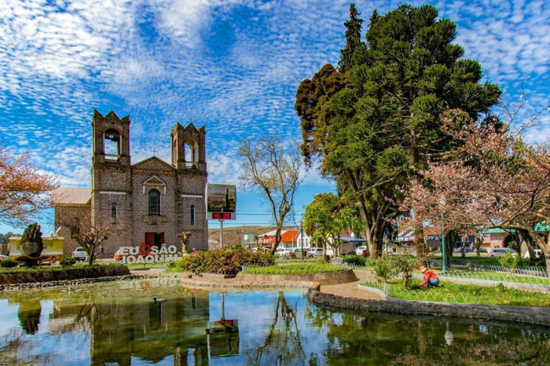 Foto da Praça Central de São Joaquim. No centro da foto, a Igreja Matriz, construída em pedras marrons e com duas torres. À frente, um totem escrito "Eu amo São Joaquim". Mais à frente ainda, um lado. Ao lado direito, algumas árvores. 