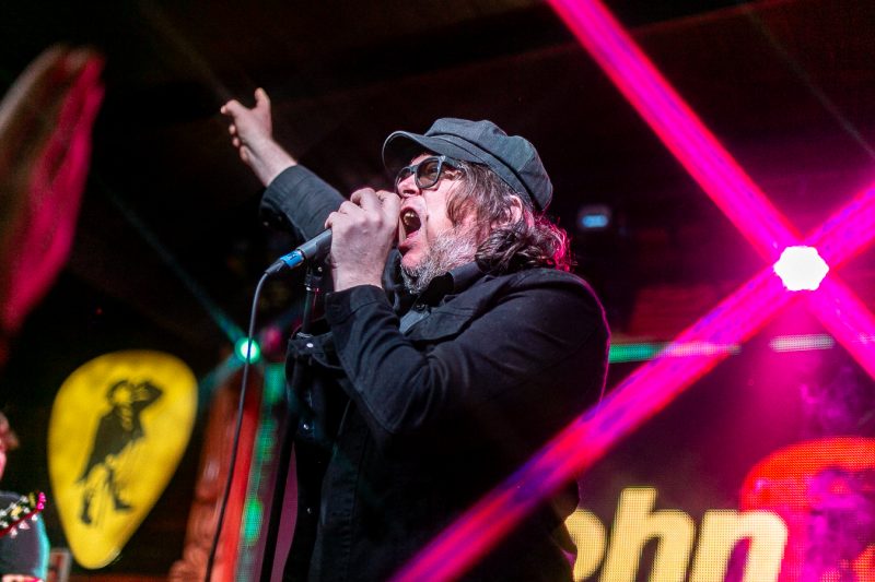 Foto de Beto Bruno no palco do John Bull, em Florianópolis. Beto é um homem branco, com cabelo castanho na altura do queixo e barba grisalha. Ele usa óculos, um paletó e uma boina preta. Com a mão esquerda ele segura o microfone e a mão direita está esticada para o alto. Ao fundo aparecem algumas logomarcas desfocadas. Uma luz rosada aparece do lado direito do artista. 