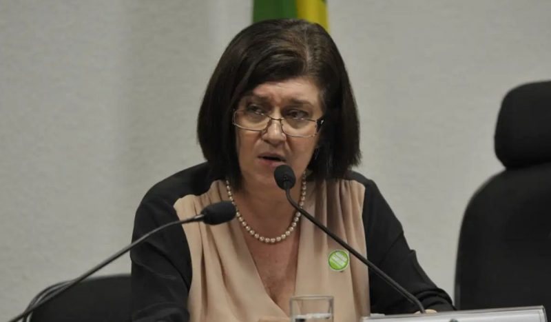 foto mostra Magda Chambriard séria enquanto fala em dois microfones, ela usa óculos, cabelos curtos e escuros, um colar de pérolas e uma blusa de mangas compridas bege e preta