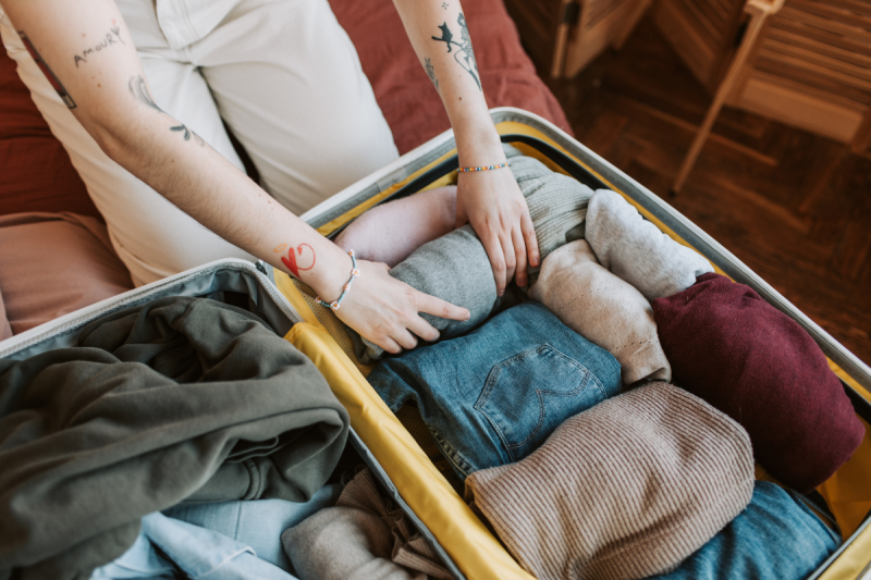 Foto ilustrativa de uma mulher jovem montando uma mala de viagem. Na foto, aparecem apenas seus braços e mãos e parte das pernas. Ela enrola uma blusa e coloca dentro da mala, que já conta com outras peças de roupa. 