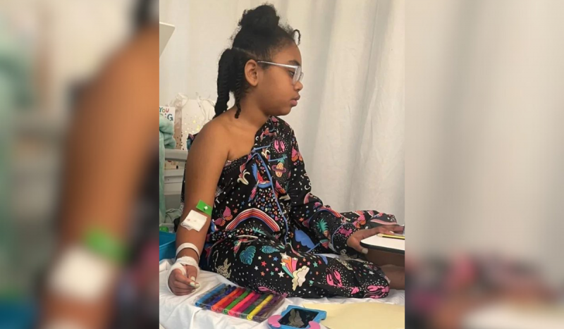 Criança com roupa preta estampada sentada em maca de hospital com acesso venoso no braço esquerdo