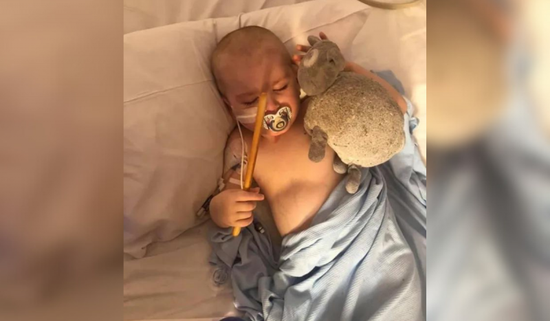 Bebê careca deitado em cama de hospital dormindo. Por cima dele um lençol azul claro. Em uma mão ele segura uma espécie de varinha amarela e na outra um urso de pelúcia