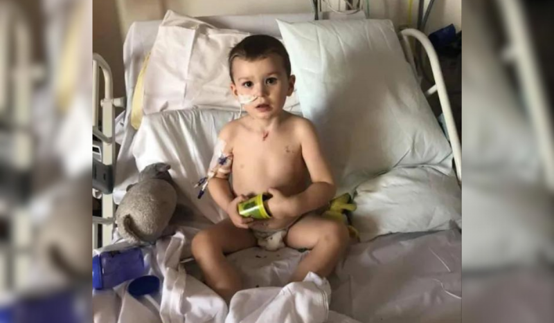 Menino branco usando tubos no nariz sentado em cama de hospital enquanto enfia a mão dentro de um copo verde de plástico 