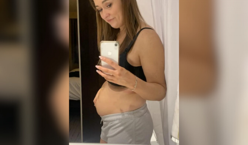 Mulher tirando foto no espelho mostrando barriga inchada. Ela usa top preto e short cinza