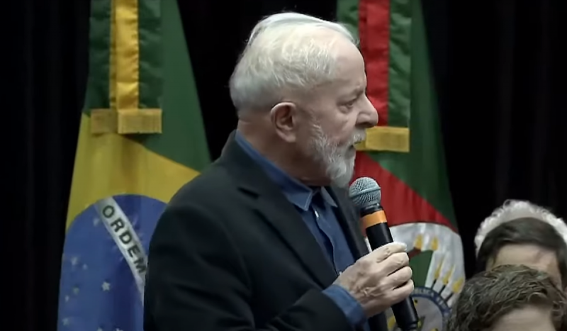 Presidente Lula com microfone na mão. Foto mostra homem branco idoso com microfone nas mãos em frente à bandeiras do Brasil e do Rio Grande do Sul
