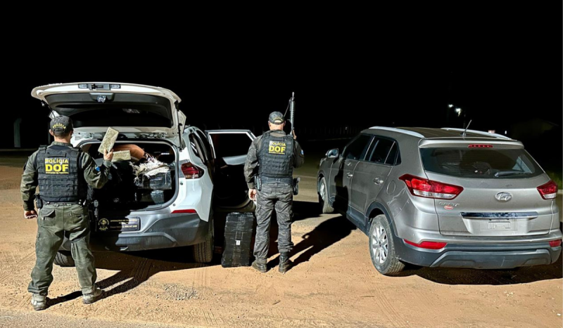 Foto mostra dois policiais de costas ao lado de dois carros, um cinza e um branco