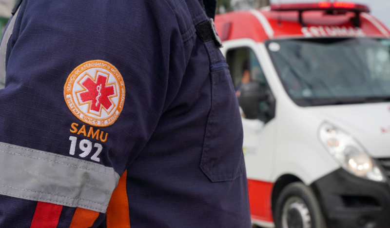 pessoa com a farda do samu e a imagem do brasão do samu no braço e ao fundo uma ambulância do samu equipe que atendeu vítima após se engasgar com comida