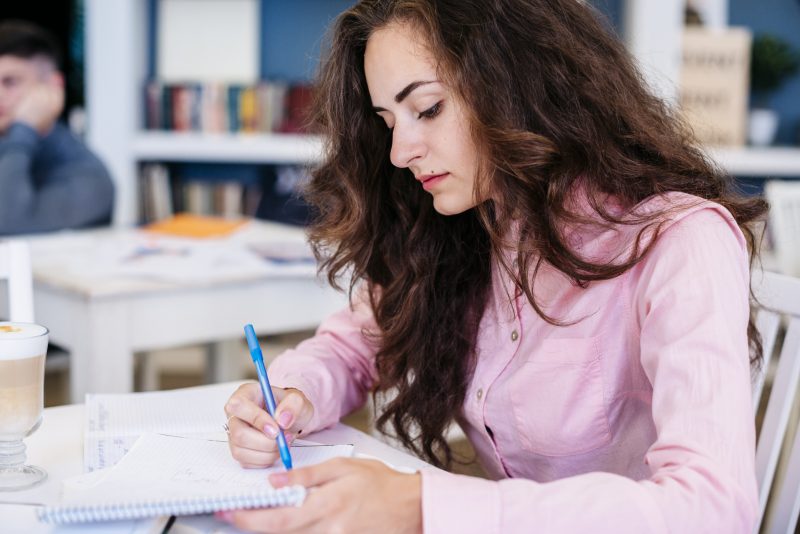 Mulher branca de camisa social rosa e cabelos longos e castanhos escuros escrevendo em um caderno com uma caneta azul claro