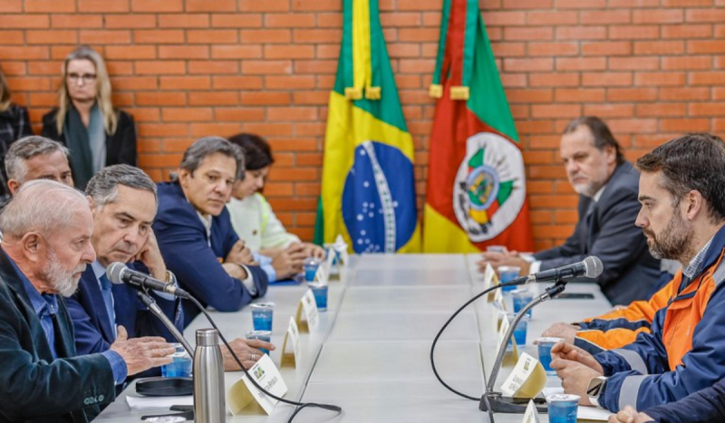 Mesa com vários homens de terno sentados discutindo. Atrás uma parede de tijolos marrons e uma mulher loira de óculos aparece em pé ao lado de duas bandeiras, uma do Brasil e outra do Rio Grande do Sul. Foto é utilizada em matéria sobre pagamento de pix ao público afetado por enchentes no Rio Grande do Sul