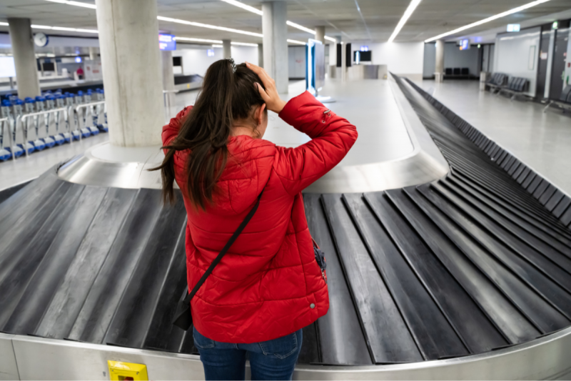 Foto ilustrativa. Uma mulher jovem, de pele clara, com cabelos longos, pretos e amarrados, fica com as duas mãos sobre a cabeça em frente à esteira de bagagens de um aeroporto, sugerindo que ela não encontrou sua mala. Ela está de costas para a foto e veste uma jaqueta vermelha e calça jeans. 