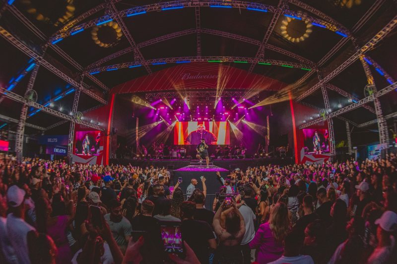 Foto do ambiente da casa de shows em Florianópolis, Stage Music Park. No centro da foto há uma passarela com um cadeira, onde está sentado o cantor Tiee. Ele é um homem negro com sobrepeso, usa uma camiseta, uma bermuda preta e um boné pretos. Ao fundo dele, o palco com luzes coloridas em amarelo e roxo. À frente e nas laterais há uma multidão de pessoas. 