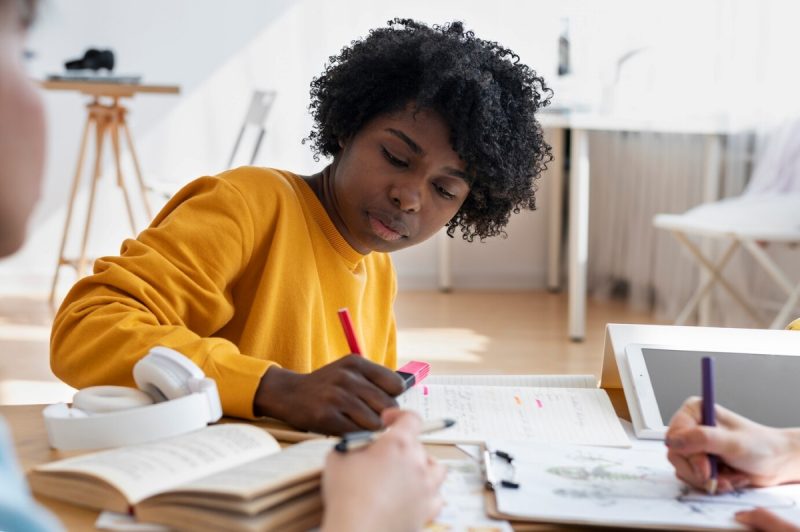 mulher negra de cabeços curtos e cacheados, ela está de moletom amarelo, sentada em uma mesa com outras duas pessoas que só aparecem as mãos, na mesa estão vários livros e cadernos e um fone de ouvido, representando pessoas inteligentes estudando, a mulher está com um lápis vermelho na mão e escrevendo em um dos cadernos, ela tem uma expressão concentrada