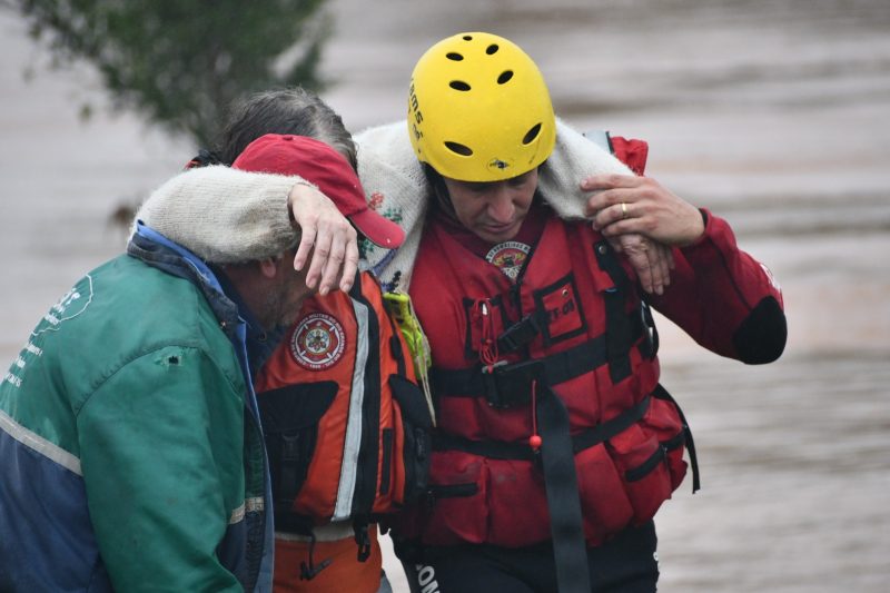 foto mostra mulher sendo carregada por bombeiro e voluntário em meio às chuvas que atingem o Rio Grande do Sul, cidades em calamidade terão prazo para entrega do imposto de renda adiado