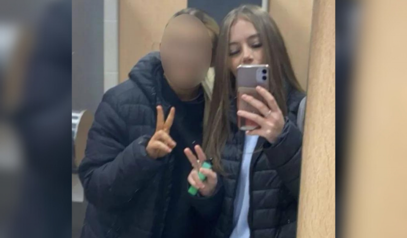 Foto mostra menina branca de cabelos loiros e compridos tirando foto no espelho segurando vape. Menino com cara borrada posa ao lado dela usando casaco preto