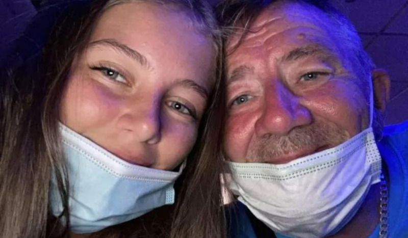 Foto mostra adolescente e pai dela usando máscara cobrindo apenas parte do queixo. Ambos são brancos de olhos verdes claros. Eles estão sorrindo em selfie.