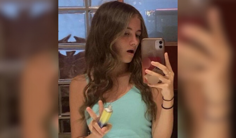 Foto mostra menina branca e loira de blusa azul tirando foto no espelho