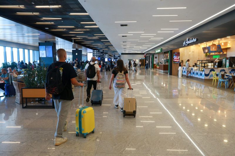 Foto do saguão de embarque do Aeroporto de Florianópolis. Três pessoas - dois homens e uma mulher estão no centro da foto com malas de rodinhas. Estão de costas para foto. Do lado direito, lojas e restaurantes. Do lado esquerdo, pessoas estão sentadas em poltronas, ao lado de grandes janelas de vidro. 