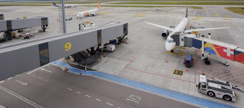 Foto com vista de cima e lateral de parte da pista do Aeroporto de Florianópolis. À esquerda dois corredores cobertos por onde passam os passageiros até chegar ao avião. Ao fundo, um avião da Gol. À direita da imagem, outro avião. 