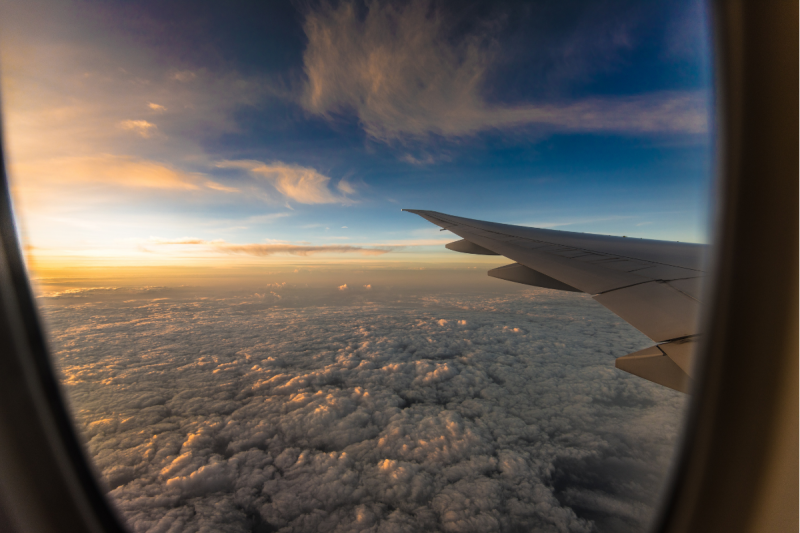 Foto da imagem de um céu azul repleto de nuvens visto a partir da janela de um avião. As bordas da janela aparecem na foto, como também uma das asas e muitas nuvens abaixo. No horizonte, há raios de sol. 