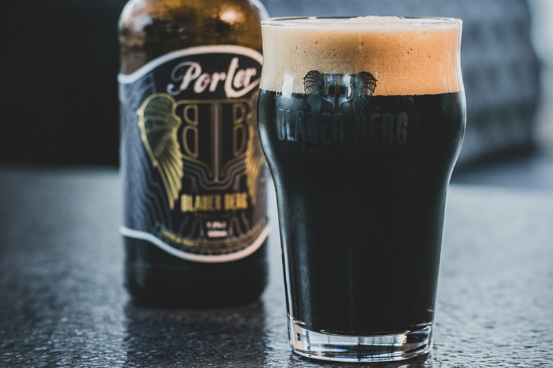 Blauer Berg Foto de um copo preenchido pela cerveja escura Porter, com uma espessa camada de espuma. Atrás do copo e desfocada, a garrafa da cerveja com a logomarca da Blauer Berg.