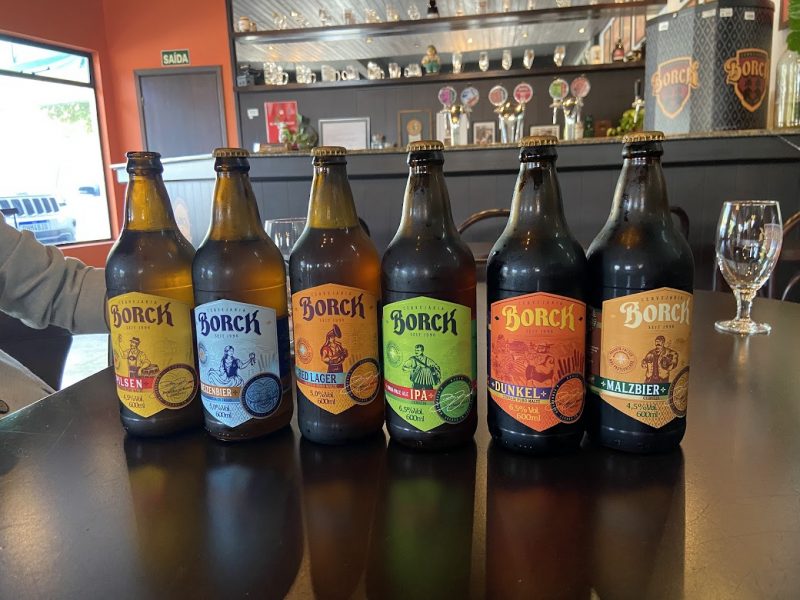 Borck Foto de seis garrafas de vidro de diferentes cervejas artesanais produzidas pela cervejaria Borck, em Timbó. As garrafas têm rótulos diferentes e estão sobre uma mesa de madeira. Atrás delas há um copo de vidro e ao fundo, um balcão de bar. O ambiente é o bar da fábrica da cervejaria.