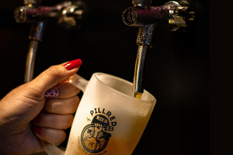 Pillred Bier Foto da mão de uma mulher com unhas vermelhas abastecendo um copo com aspecto de congelado em uma torneira de chope. O copo tem a logomarca da cervejaria Pillred Bier, de Timbó.