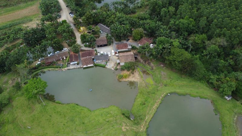 Foto aérea da propriedade família Hattenhauer. No centro da foto aparecem duas grandes lagoas. Ao redor há um gramado e algumas casas. Na parte superior e direita da foto aparecem várias árvores. No canto mais esquerdo aprece uma estrada de chão. 