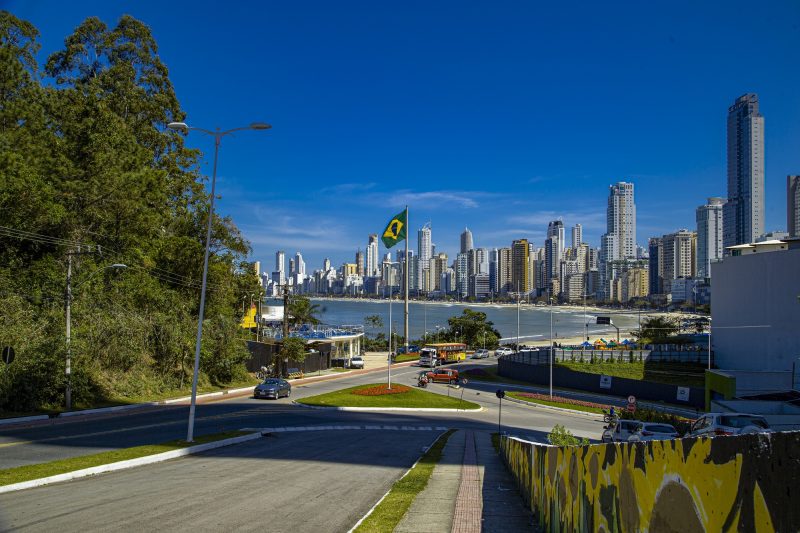 Foto do final da descida da Estrada da Rainha em direção à Praia Central de Balneário Camboriú. No centro, as duas pistas de trânsito. Do lado direito, aparece uma parte de um muro grafitado. Do lado esquerdo, parte da vegetação do Morro da Rainha. À frente, o céu azul, o mar e a orla da Praia Central cercada por altos prédios. 