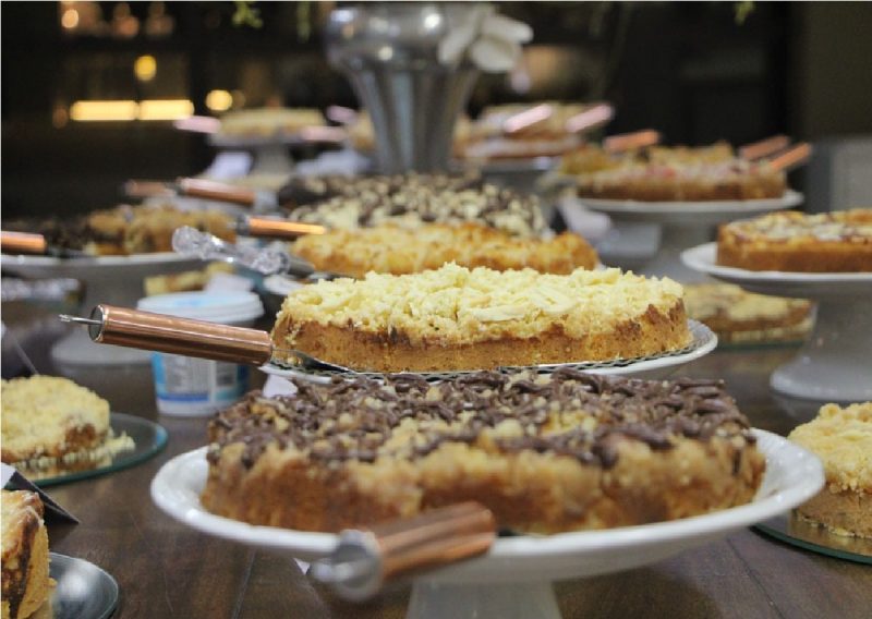Imagem mostra diversas cucas, iguaria típica de SC, em uma mesa bem arrumada. Todas elas contam com uma espátula e são diversas coberturas nas cores marrom e branco