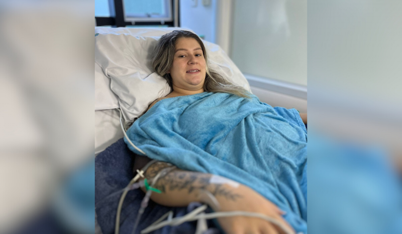 Foto mostra paciente sorrindo deitada em cama de hospital enquanto está ligada a aparelhos no braço direito. Mulher é branca, loira, usa aparelho nos dentes e tem cabelos tamanho médio. Ela veste um roupão azul celeste. Mulher teve o feto de 24 semanas operado ainda no útero