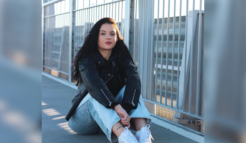 La foto muestra a una niña blanca con ojos celestes sentada en el suelo.  Viste una chaqueta de cuero negra y jeans azul claro, tiene cabello largo y negro y tiene una expresión seria en las fotos.